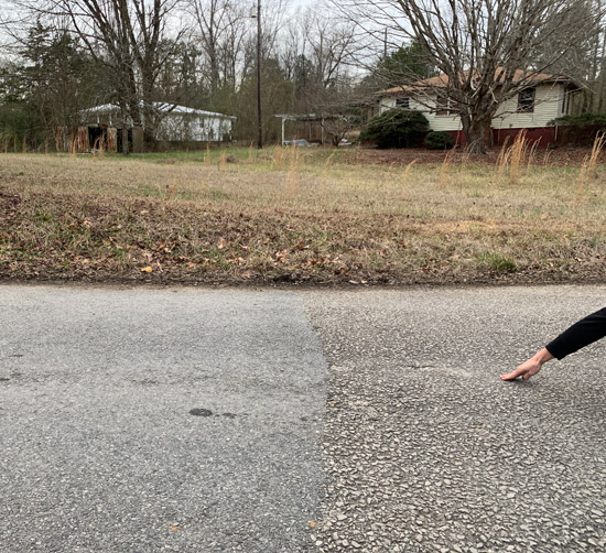 Photo of a man's arm/hand indicating the location on the road. In the background is a dried grass front yard and a cream colored house one-storey house with a reddish roof.